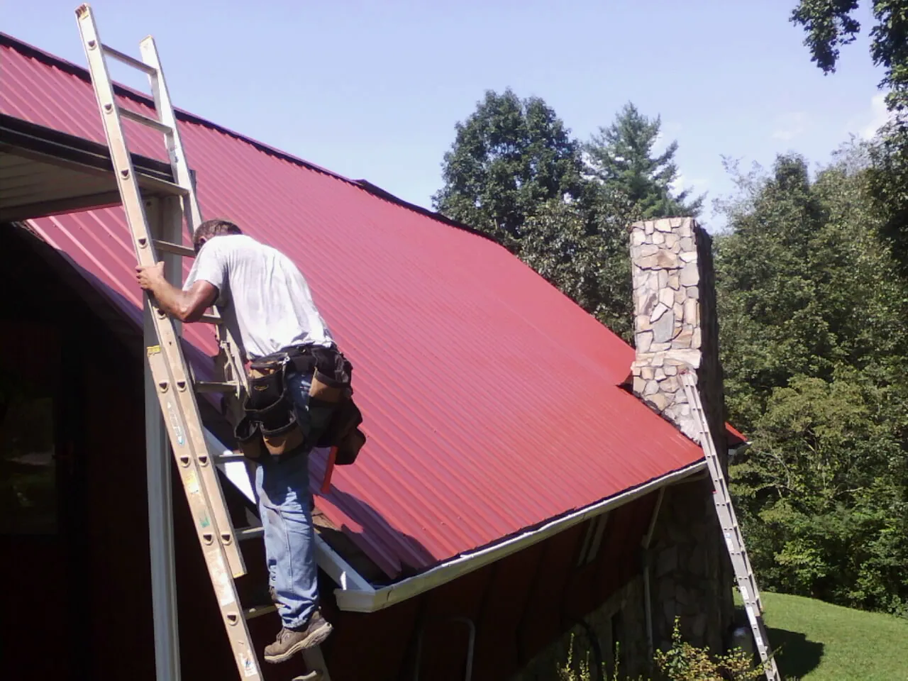 Roofer on a ladder beside a red metal roof and stone chimney — Soffit & Fascia Repair by Roofing Brothers in New Britain